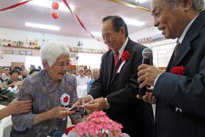 トシさんはすっくと立ち上がって、儀間会長から記念品を受け取った。右が宮城あきらさん