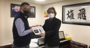 学校法人力行会の田中総務部長から３冊を受け取る加藤代表